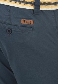 Shorts en tissu bleu-gris avec une surface texturée, comportant une étiquette en cuir marron, une fermeture par bouton et une taille rayée contrastante.