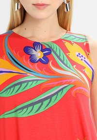 Červená halenková bez rukávů s výraznými květinovými a listovými vzory v modré, zelené, žluté a růžové barvě. Hladká látka s kulatým výstřihem.