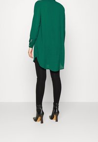 Groene tuniek met lange mouwen en zijsplitsen, zwarte legging en zwarte enkellaarsjes met hak. Gladde stof en een losse pasvorm.