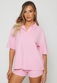 Frau mit langen, welligen Haaren, die ein kurzärmeliges, pink-weißer gestreiftes Hemd mit Kragen und passende Shorts trägt, steht vor einem einfarbigen Hintergrund.
