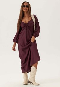 Lungo vestito bordeaux con parte frontale arricciata e maniche voluminose, indossato con stivali da cowboy beige. Tessuto morbido e orlo asimmetrico.