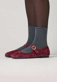Zapatos Mary Jane de ante rojo con un hebilla dorada redonda, combinados con calcetas de rayas grises, mostrados desde el tobillo hacia abajo sobre un fondo neutro.