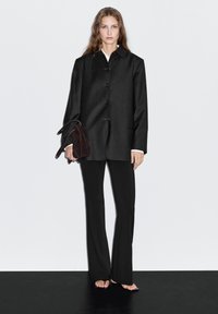 Black wool-blend jacket with a button-up design, paired with black flared pants. The model holds a small dark brown suede bag.