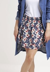 Woman wearing a blue floral skirt, white top, and a denim jacket with rolled sleeves, standing with legs crossed.