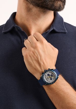 Kék gumiszíjas óra ezüst számlappal, három segédszámlappal és kerek tokkal. Texturált sötétkék galléros póló a háttérben.