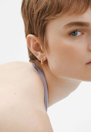 Pendiente de aro dorado con un diseño retorcido, llevado en la oreja. La persona tiene el cabello corto y lleva una blusa de color púrpura claro.