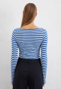 Long-sleeve striped top in blue and white, featuring a boat neckline, fitted design, and a smooth, soft fabric. Paired with dark trousers.