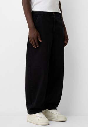 Person iført løstsiddende sorte bukser, hvide sneakers og en hvid skjorte, stående mod en ensartet lys baggrund.