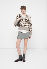 Pull en tricot avec une base beige et un motif géométrique marron, chemise blanche en dessous, jupe plissée grise, chaussettes grises et chaussures bordeaux.