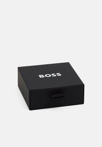Scatola rettangolare nera con una superficie liscia, dotata di una maniglia a strappo e del logo "BOSS" stampato in bianco sul lato anteriore.