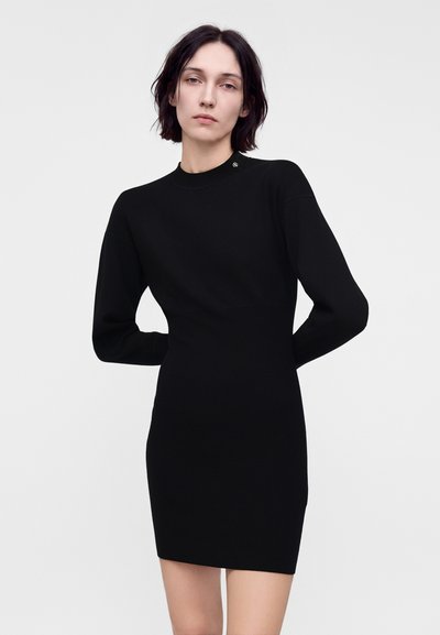 Femme aux cheveux courts et foncés portant une robe noire cintrée à manches longues, debout devant un fond clair uni.
