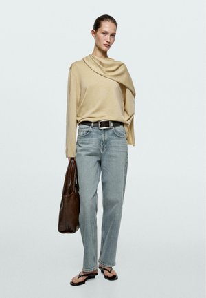 Donna in piedi che indossa un top beige drappeggiato, jeans azzurri, cintura nera, sandali neri, con una grande borsa tote in pelle marrone.