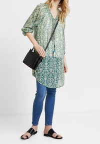 Genomskinlig grön tunika med vitt blommönster, rund halsringning och trekvartslånga ärmar, i kombination med blå slim-fit jeans och svarta sandaler.