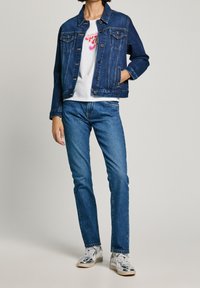 Giacca di jeans blu scuro con chiusura a bottoni, due tasche sul petto e cuciture a contrasto, abbinata a jeans azzurri e sneakers argento.