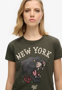 T-shirt vert olive avec un col ras du cou, présentant un graphique d'un panthère rugissante et d'une rose, avec le texte "NEW YORK" au-dessus en couleur claire.