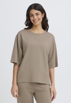 Ung kvinde med mørkt hår smiler, iført beige oversized kortærmet top og matchende bukser, stående foran en ensfarvet baggrund.