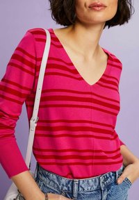 Rosa V-Ausschnitt-Pullover mit roten horizontalen Streifen, aus Strickmaterial gefertigt. Kombiniert mit hoch taillierter Jeans und einer weißen Schultertasche.