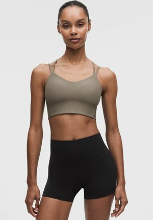 Mujer atlética de pie, con un sujetador deportivo oliva acanalado con tirantes finos y pantalones cortos de entrenamiento negros de cintura alta.