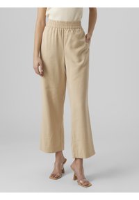Pantalon beige à jambes larges avec une taille élastiquée et des poches latérales. Tissu léger avec une texture lisse, associé à des sandales à talons transparents.