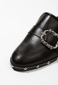 Chaussure noire en cuir à enfiler avec un bout pointu, une boucle argentée ornée et des clous le long du bord de la semelle. Texture lisse, design raffiné.