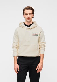 Φούτερ με κουκούλα σε κρεμ απόχρωση, με τσέπη καγκουρό, ρυθμιζόμενο κορδόνι στην κουκούλα και κεντημένο λογότυπο σε κόκκινο και μπλε. Φοριέται με σκούρο παντελόνι.