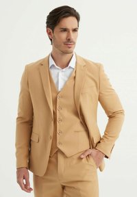 Costume trois pièces beige avec une veste à un boutonnage, des revers crantés et un gilet à cinq boutons. Chemise habillée bleu clair en dessous.