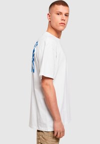 White cotton t-shirt with loose fit, featuring blue text on the left side. Round neckline and short sleeves. Plain design without patterns.
