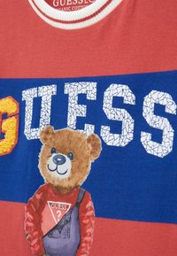 Red and blue t-shirt made of organic cotton, featuring a textured "GUESS" logo and a cartoon bear wearing a red shirt and blue overalls.