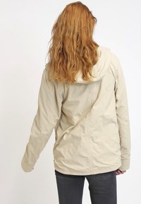 Lysebeige jakke med hætte, der har en blød tekstur, lange ærmer og synlige syningsdetaljer, båret baglæns af en person.