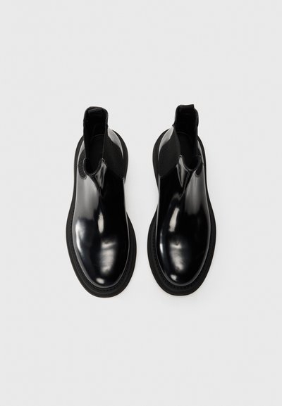 Botas Chelsea negras brillantes con un acabado suave, paneles laterales elásticos y una suela de goma negra texturizada. Diseño minimalista.
