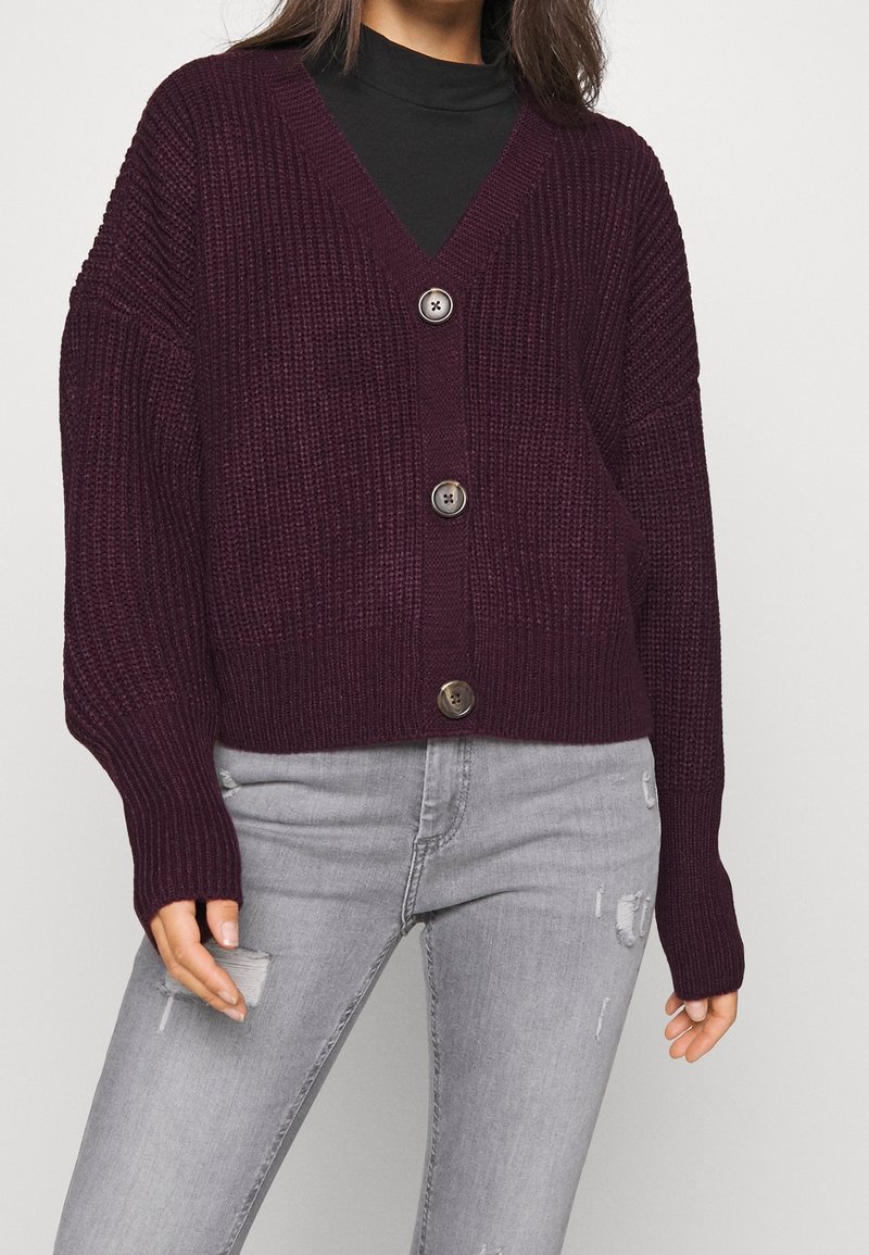 Femme portant un cardigan en maille épaisse violet foncé boutonné par-dessus un t-shirt noir avec un jean skinny gris clair déchiré.