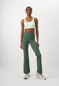 Una canotta sportiva verde chiaro abbinata a leggings svasati verde scuro a vita alta e scarpe da ginnastica grigio chiaro. Tessuto liscio e design aderente.
