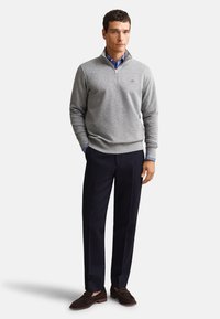 Homme portant un sweat-shirt gris à fermeture éclair au quart sur une chemise rayée bleue, un pantalon noir et des mocassins marron foncé, debout sur fond blanc.