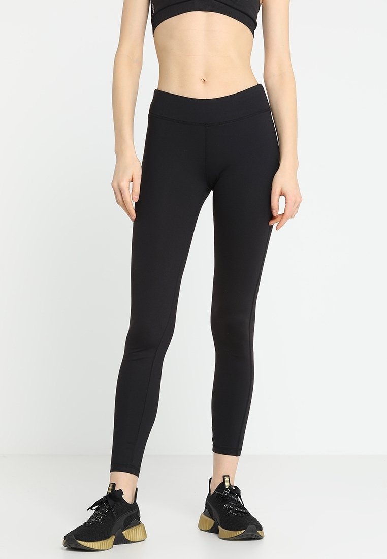 Nainen, joka on pukeutunut mustiin korkeavyötäröisiin leggings-housuihin ja mustiin urheilukenkiin, joissa on vaaleanbeige pohja, seisoo valkoista taustaa vasten.