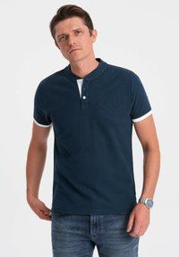 Námořnická modrá polo košile s texturovaným designem, bílým límcem a krátkým rukávem. Obsahuje dva knoflíky na léga. Volný střih.