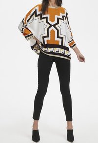 Tunica ampia con motivi geometrici arancioni, bianchi e neri e accenti metallici, abbinata a pantaloni neri aderenti e tacchi neri.