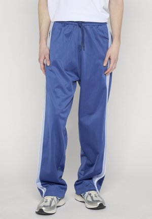 Person trägt blaue Sporthose mit weißen Seitenstreifen und silberne Laufschuhe, steht vor einem schlichten hellen Hintergrund.
