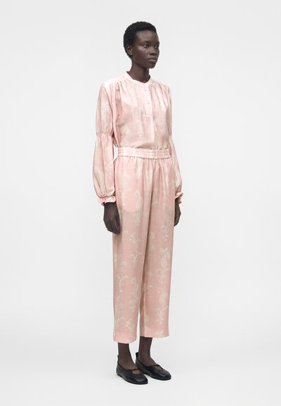 Γυναίκα που φορά μια φαρδιά, ανοιχτό ροζ πουκαμίσα με μοτίβο και ταιριαστό ψηλόμεσο παντελόνι με μαύρα επίπεδα παπούτσια, στέκεται μπροστά σε μια απλή φόντο.