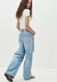 Helles blauweite Jeans mit gerissenem Saum, Fünf-Taschen-Design und einem braunen Lederpatch auf der Rückseite. Modell trägt ein enges weißes Oberteil.