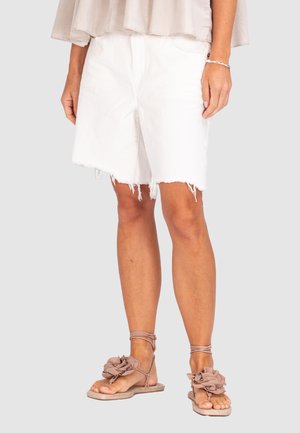 Witte denim shorts met een rafelige zoom, gecombineerd met lichtbruine sandalen met ruches. De achtergrond van de afbeelding is neutraal grijs.