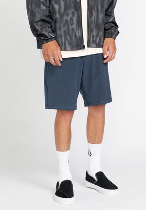 Veste légère à motifs gris foncé, avec fermeture éclair ; t-shirt blanc ; shorts bleus ; chaussettes blanches ; chaussures noires à enfiler avec semelles blanches.