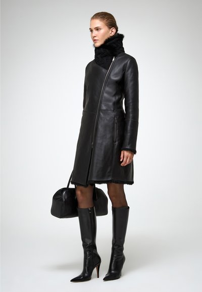 Donna che indossa un cappotto di pelle nera con colletto alto, stivali neri fino al ginocchio, che tiene in mano una borsa nera, in piedi contro uno sfondo semplice.