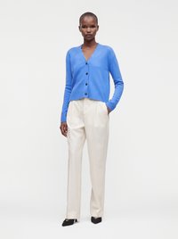 Femme portant un cardigan bleu boutonné, un pantalon blanc plissé et des talons noirs, debout avec une main dans la poche devant un fond uni.
