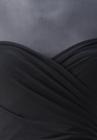 Svart kjole i stoff med teksturert overflate, med draperte detaljer over kroppen og en jevn enkel farge på den øvre delen.