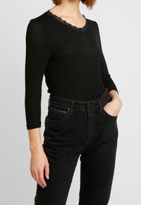 Musta pitkähihaisa paita, jossa on kihara pitsikaulus, yhdistettynä mustiin korkeavyötäröisiin farkkuihin, joissa on etutaskut ja slim fit.