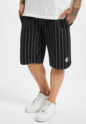 Shorts à rayures noires en tissu doux, avec des lignes verticales blanches, un logo brodé en bas et des poches latérales. Portés avec des baskets blanches.