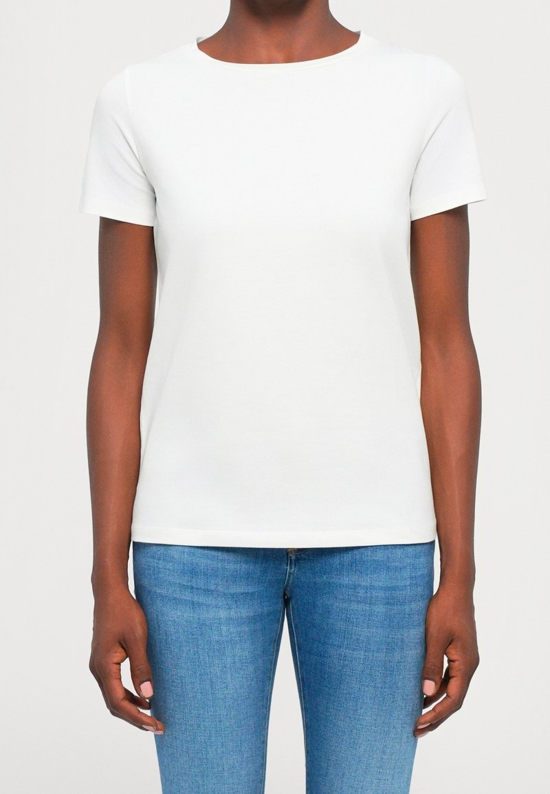Personne portant un t-shirt blanc uni à manches courtes et un jean bleu, debout devant un fond neutre, les bras détendus le long du corps.
