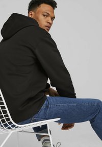 Black hoodie with a textured surface, featuring a hood and long sleeves. Worn with blue jeans, seated on a white wire chair.
