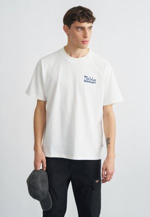 T-shirt en coton blanc avec logo "Dickies" bleu, manches courtes, coupe décontractée. Le mannequin tient une casquette gris foncé. Pantalon cargo noir visible.