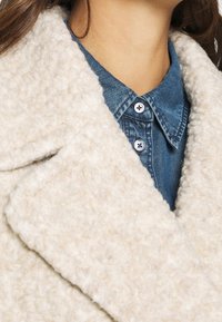 Nærbillede af en person iført en blå denim skjorte med knapper, lagdelt under en cremefarvet struktureret fleecejakke.